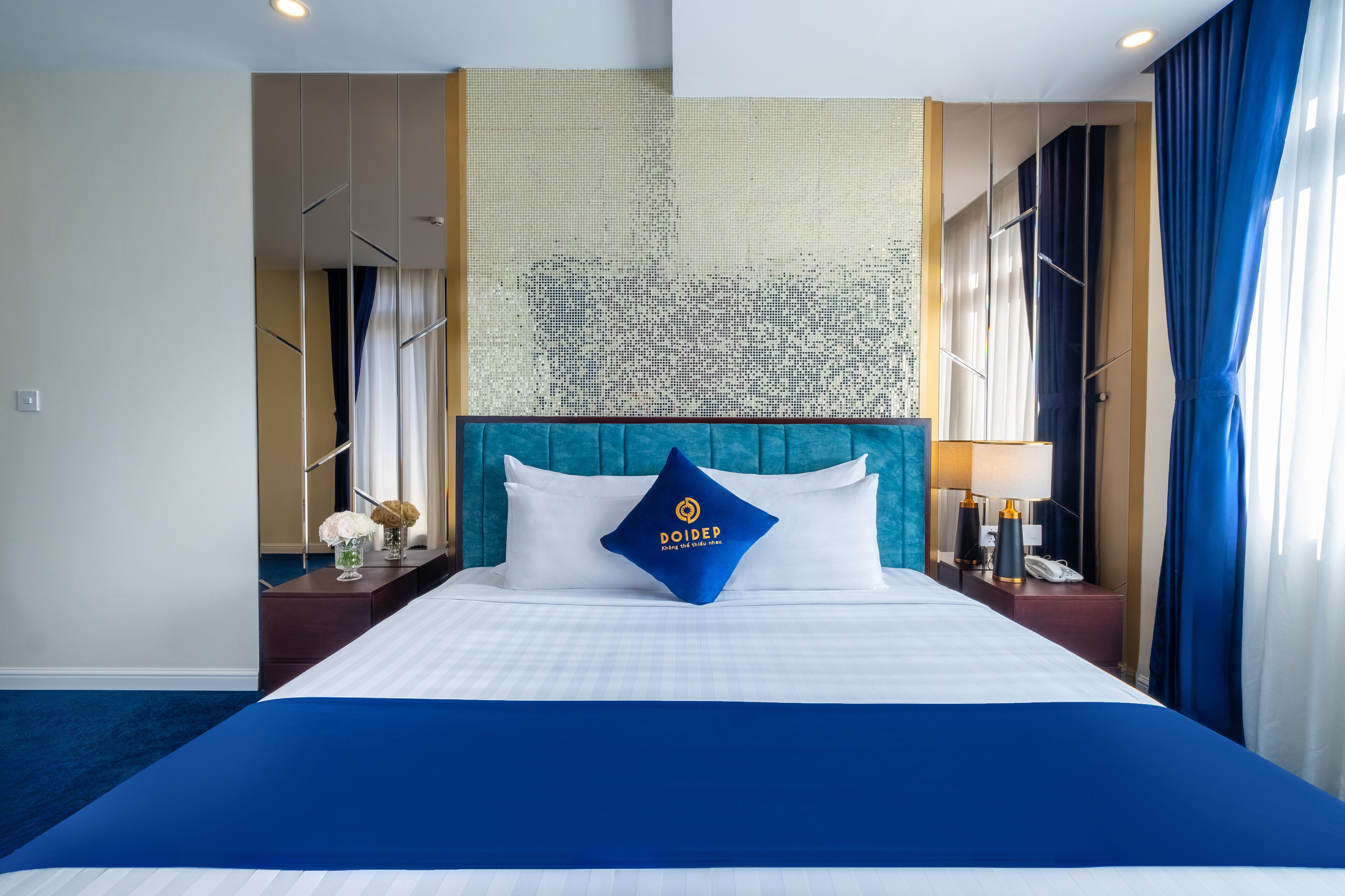A hotel room with a large bed, a headboard with a logo, a nightstand with a lamp, and a view of the city through the window.