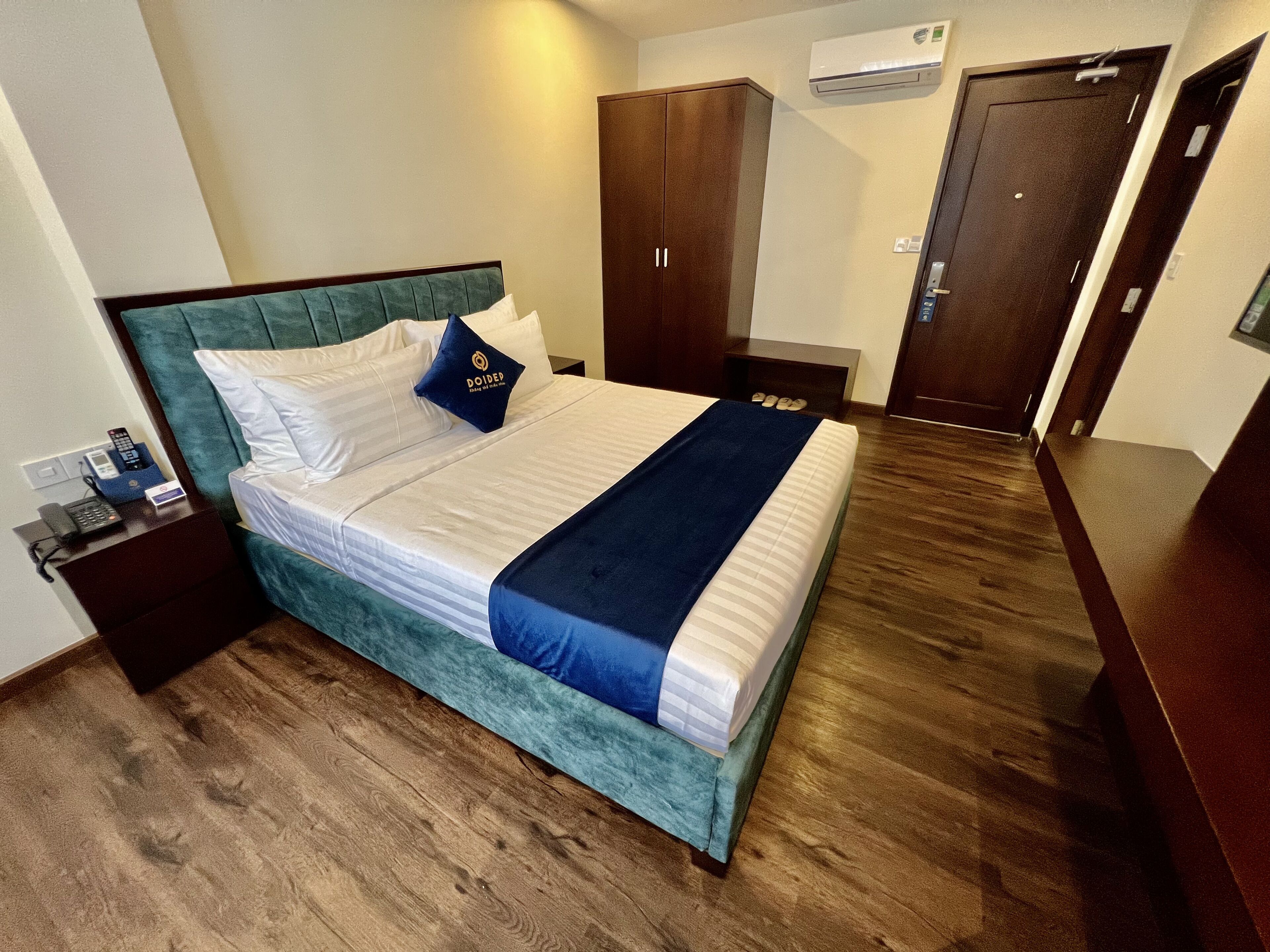 A neatly made bed with a blue blanket, a wooden headboard, and a pillow with a logo. There is a wooden wardrobe, a small bedside table with a phone, and a wall-mounted air conditioner.