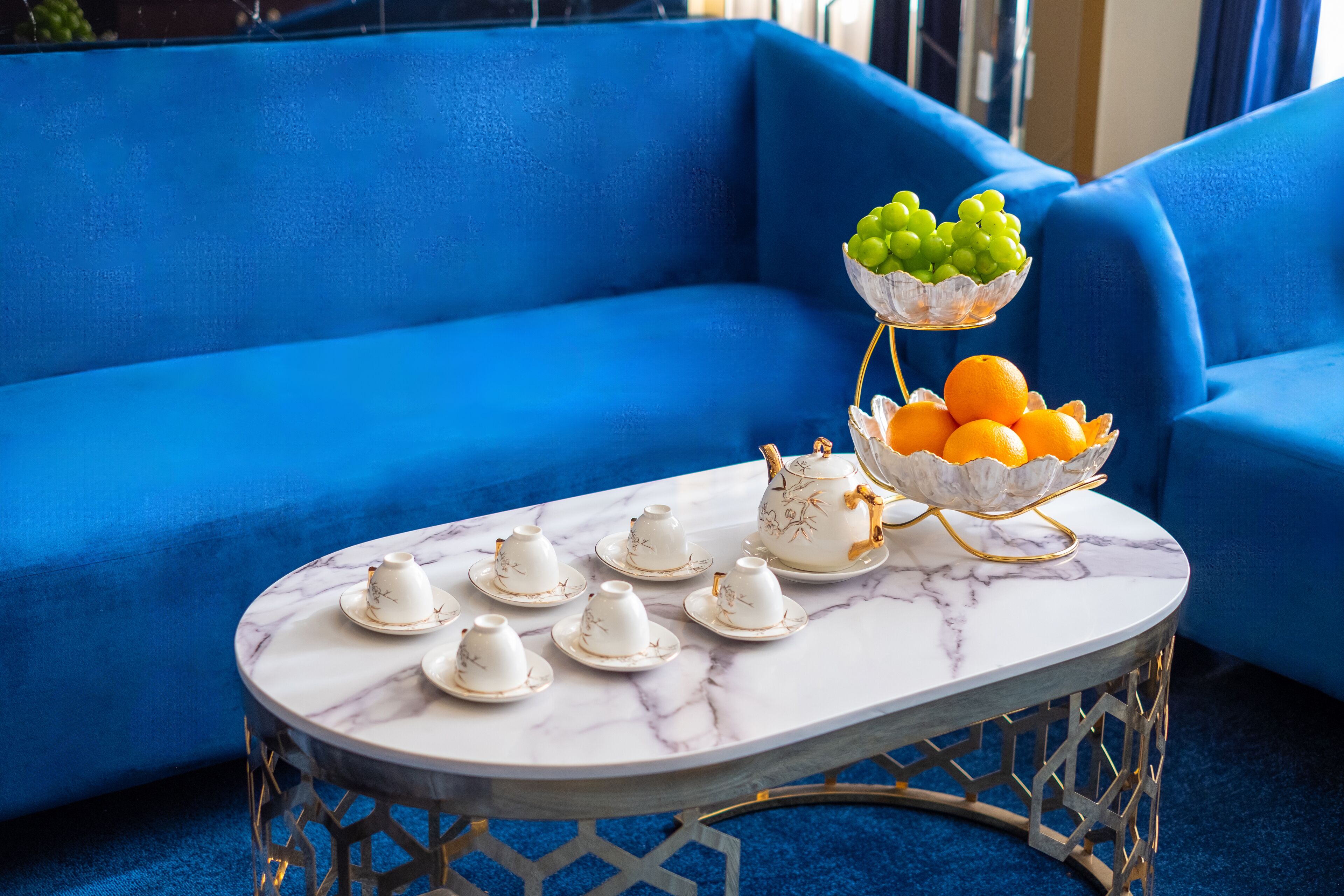 A blue sofa, a marble table with teapots and cups, and a fruit bowl with grapes and oranges.