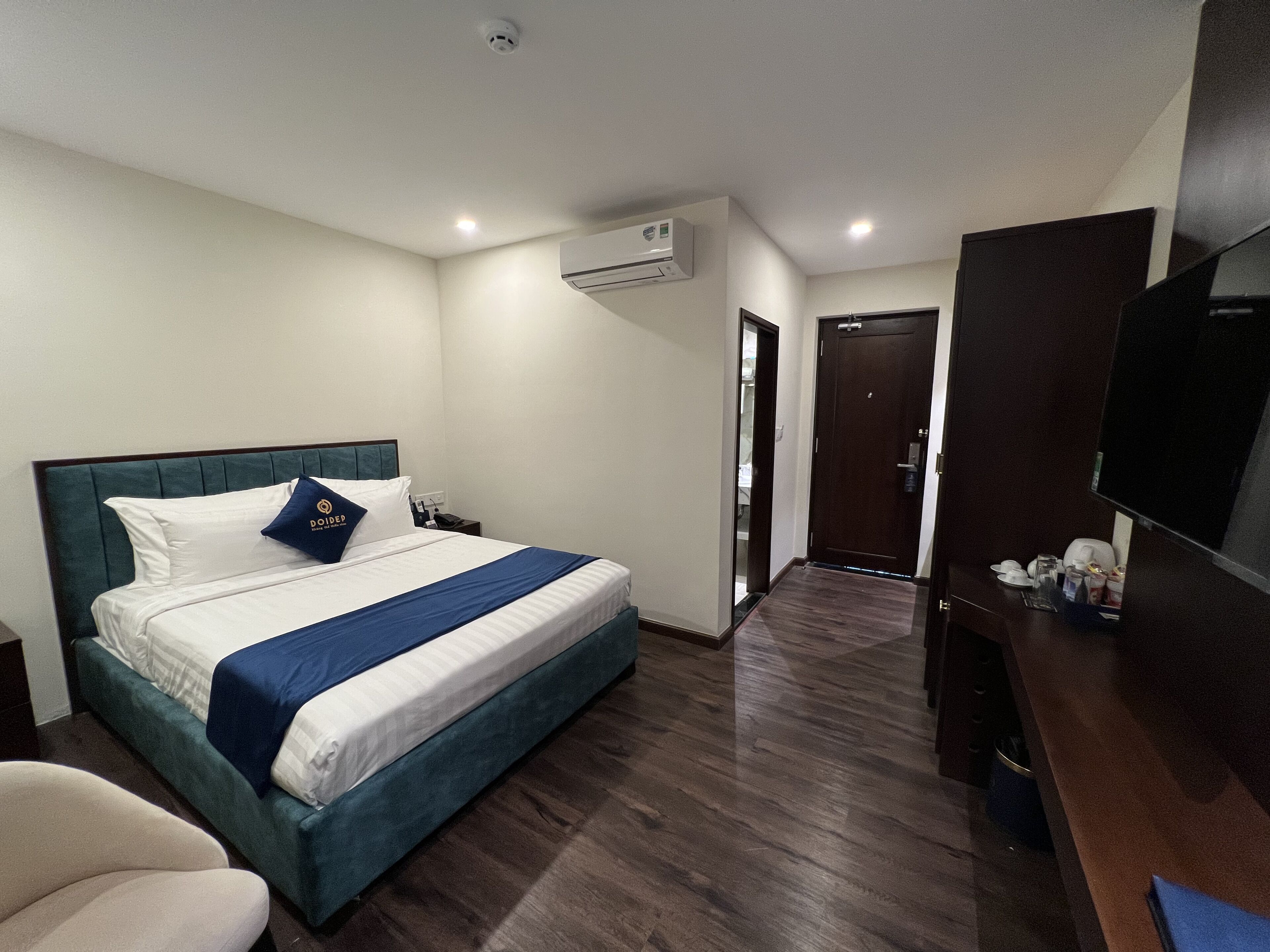A hotel room with a bed, a desk, a chair, an air conditioning unit, and a television.