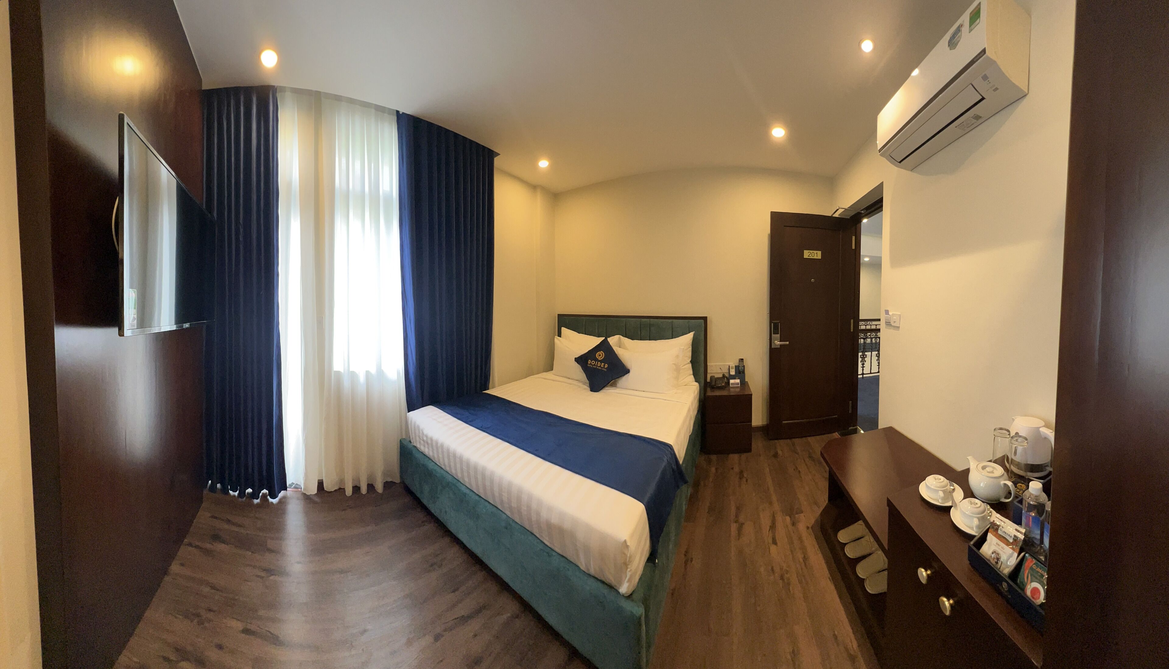 A hotel room with a bed, a bedside table with a tray of tea and coffee, a wardrobe, and an air conditioning unit.