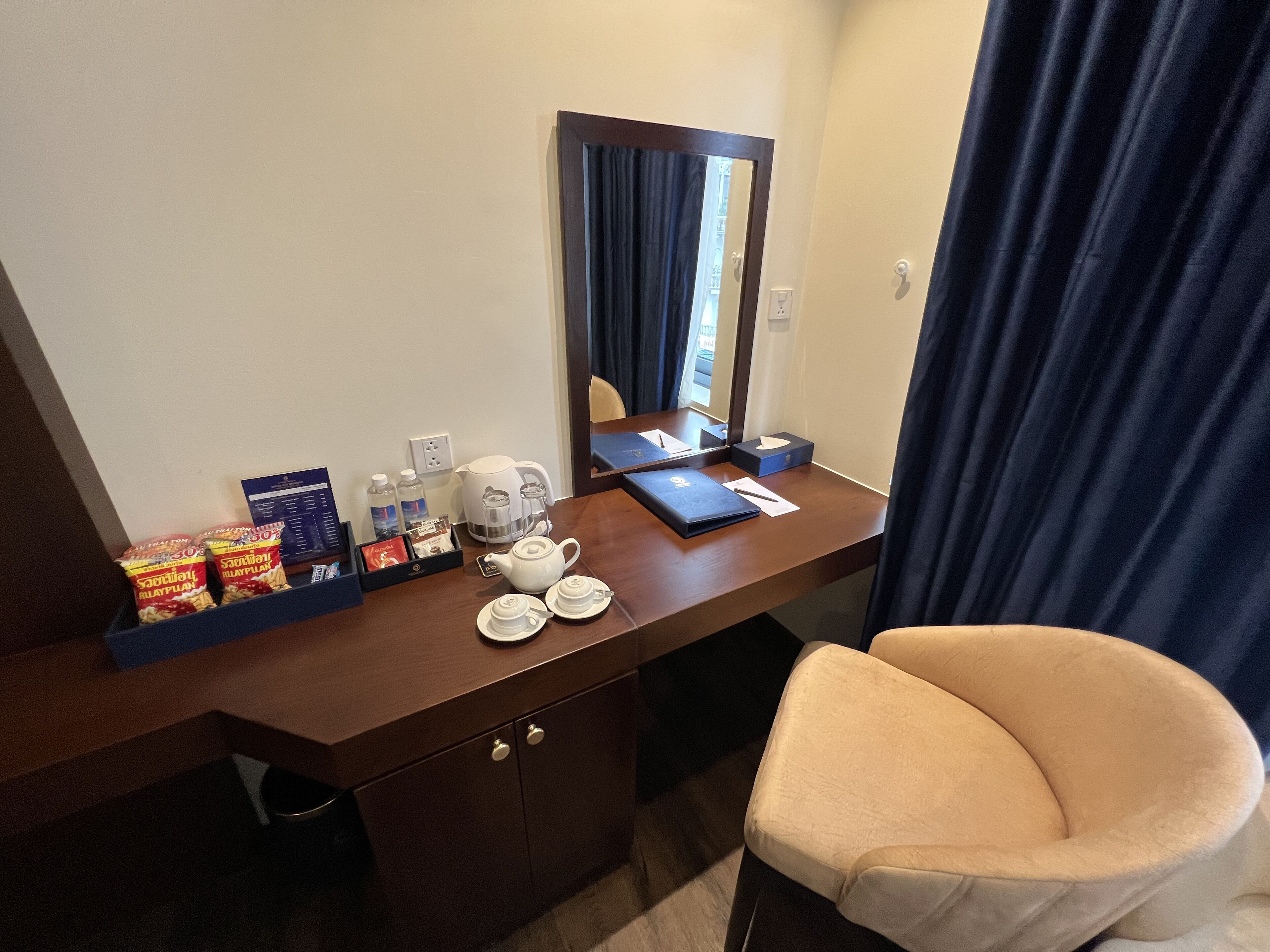 Une chambre d’hôtel avec un bureau en bois, un miroir, une chaise et une petite poubelle.