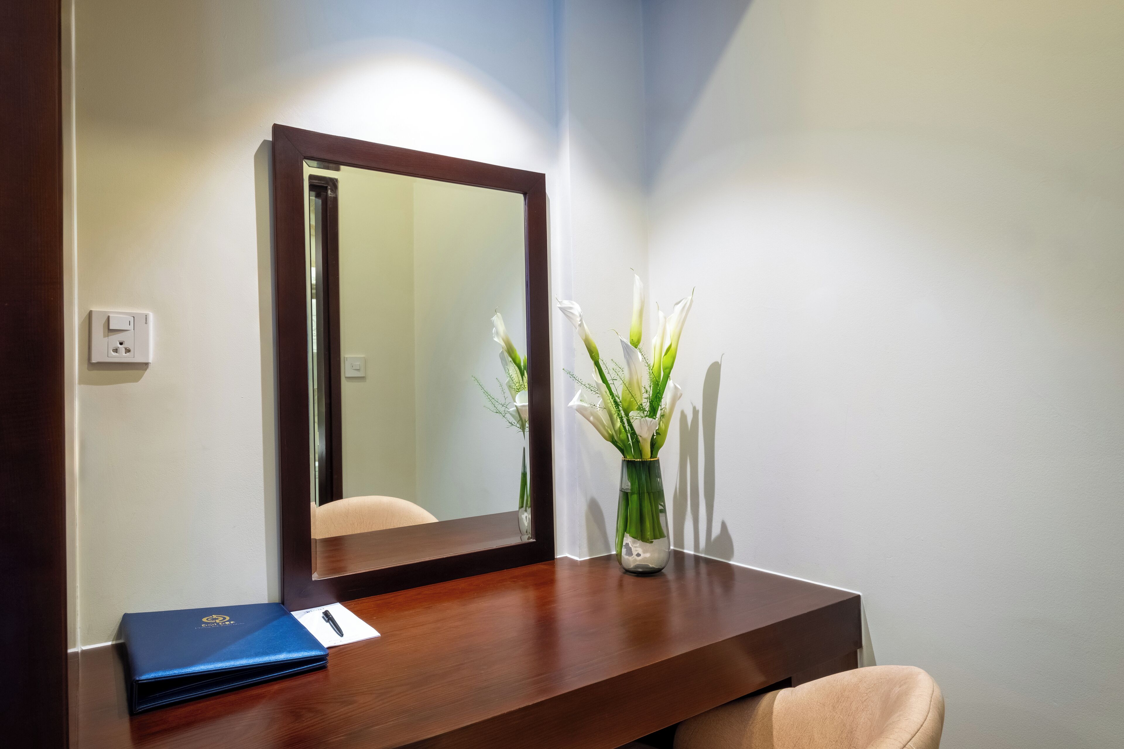 A hotel room with a wooden desk, a mirror, a vase with flowers, and a blue folder.