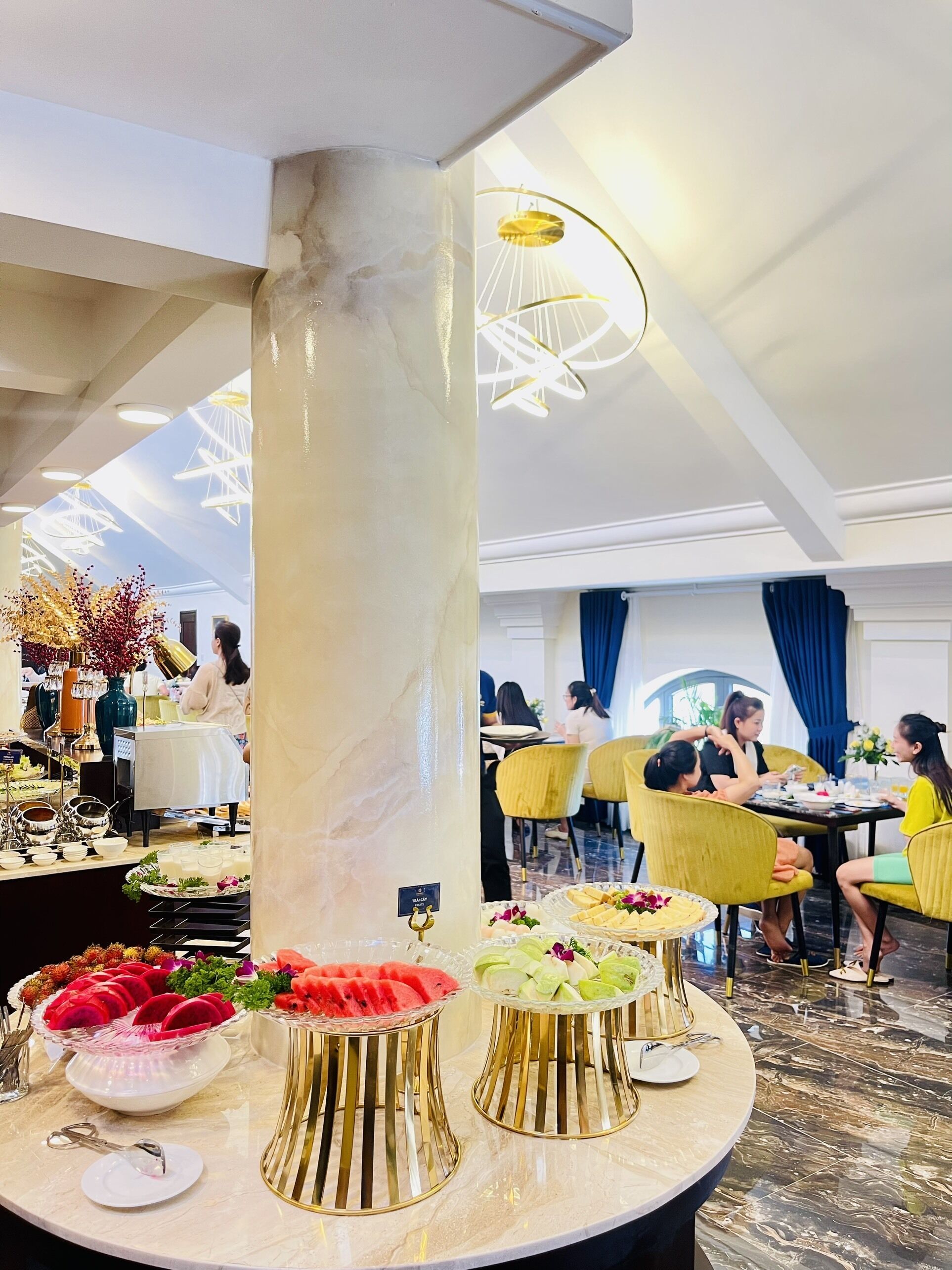 Um saguão de hotel com colunas de mármore, uma área central de serviço com comida e bebidas, e um espaço de estar com cadeiras amarelas.
