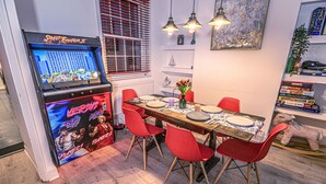 Un coin repas avec une borne d’arcade rétro, des chaises rouges, une table en bois et une bibliothèque contenant des livres et un jouet en peluche.