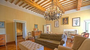 Un vaste salon doté d’un parquet, d’un grand canapé beige, d’un pouf blanc et d’un lustre. Les murs sont ornés d’œuvres d’art encadrées, et on aperçoit un coin repas au fond de la pièce.
