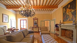Un vaste salon doté d’un plafond en bois, d’un grand lustre, d’un canapé beige, d’un pouf blanc et d’une cheminée agrémentée d’objets décoratifs.