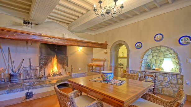 Un coin repas chaleureux avec une table en bois et des chaises en osier, une cheminée où l’on peut voir le feu brûler, et des éléments décoratifs en pierre sur les murs.