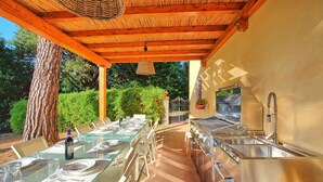 Un espace extérieur aménagé pour les repas, avec une longue table dressée pour un repas, un coin barbecue et une pergola au-dessus.