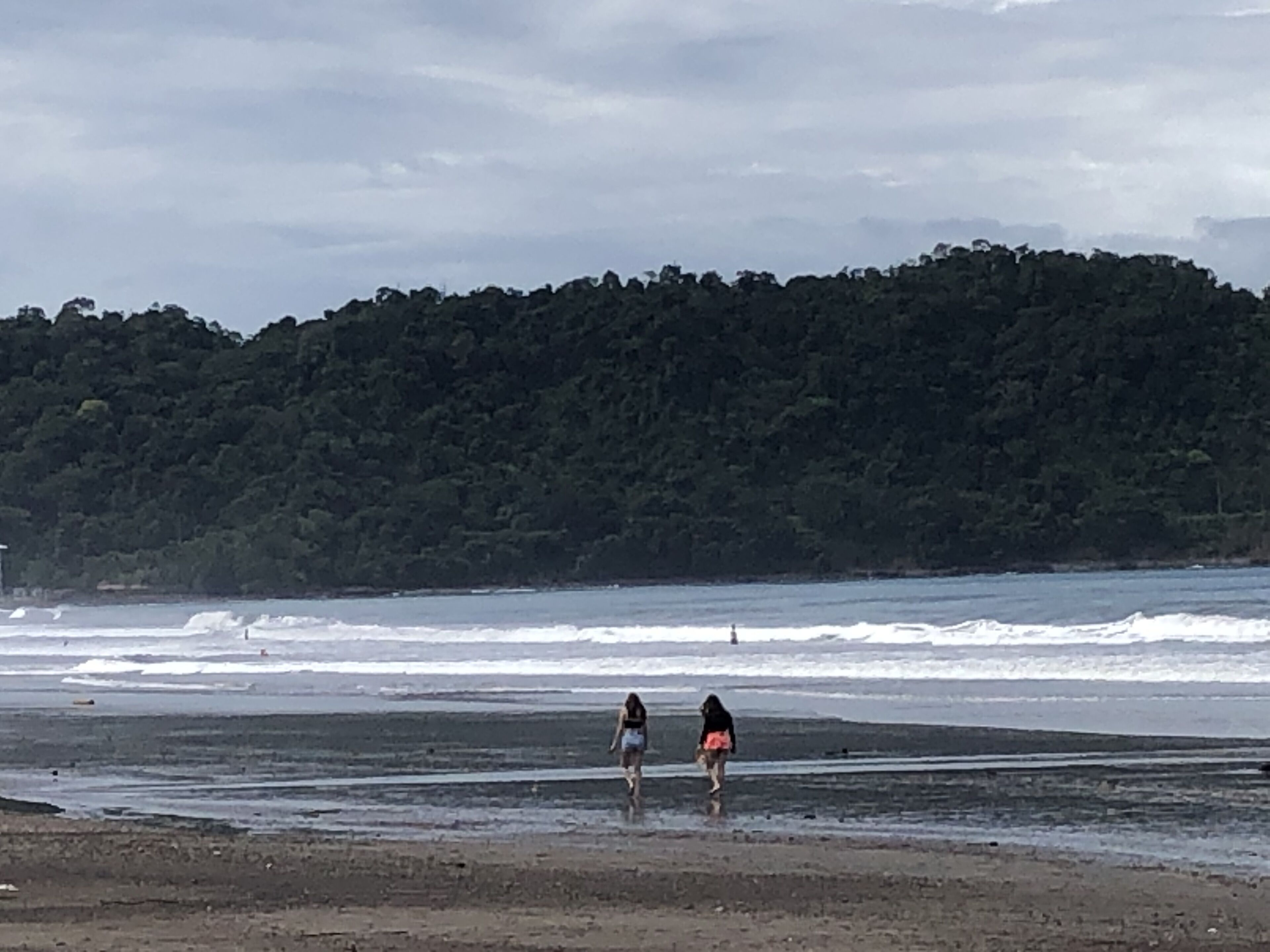 Una playa con dos personas caminando por la orilla, una colina boscosa al fondo y olas rompiendo sobre la arena.