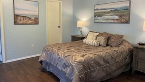 Quarto com cama grande, duas pinturas nas paredes, uma porta e mesa de cabeceira com abajur.