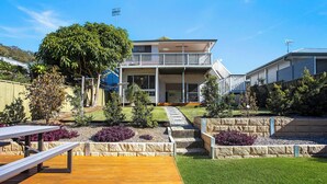 A two-story house with a balcony, a wooden deck, and a well-maintained garden with various plants and a stone retaining wall. - Renovated 3 bed Unit with Views (Umina Beach)