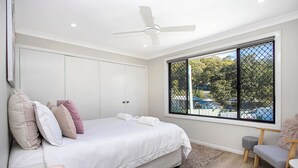 A bedroom with a large bed, a ceiling fan, a window with a view of trees and a pool, and a small seating area. - Renovated 3 bed Unit with Views (Umina Beach)
