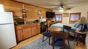 A compact kitchen with wooden cabinets, a refrigerator, a microwave, and a coffee maker. There is a dining area with a table and chairs, and a bed with a blue blanket.