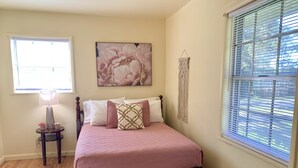Quarto com cama, mesa de cabeceira, abajur, um enfeite de macramé na parede e janela com persianas.