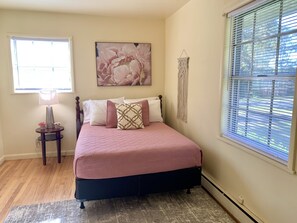A bedroom with a bed, a nightstand, a lamp, a macrame wall hanging, and a window with blinds.