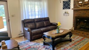 Uma sala de estar com sofá de couro marrom, mesa de centro de madeira e tapete colorido. Há uma lareira com revestimento de tijolos e objetos decorativos na mantel.