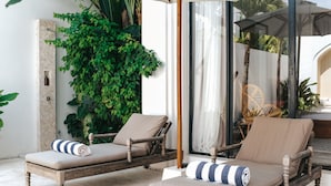 Two wooden lounge chairs with cushions and striped towels, positioned on a patio with a view of a lush green plant and a glass door leading to an interior space. - Villa M Canggu (Canggu)