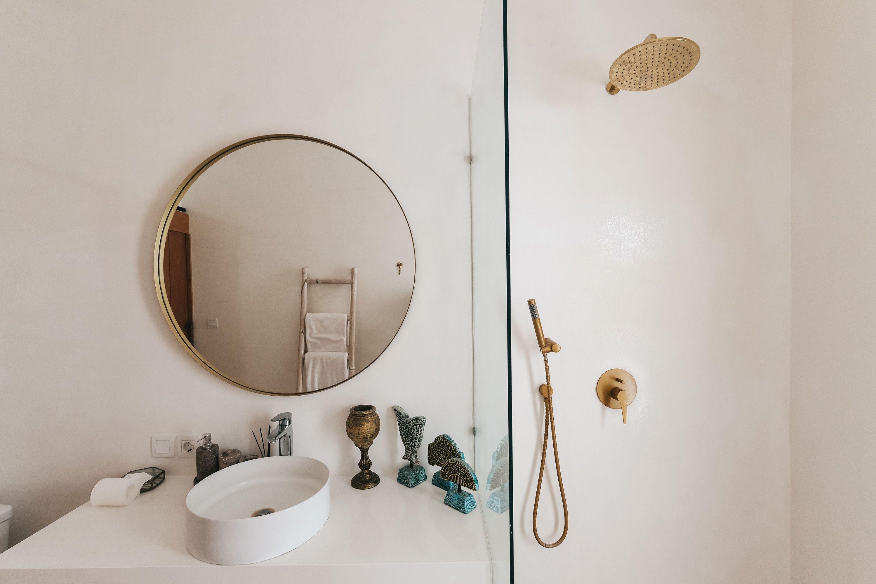 Une salle de bain avec un miroir rond, un lavabo blanc, une pomme de douche dorée et des objets d écoratifs.