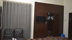 A room with a dark brown door, a small TV mounted on the wall, a grey curtain, and a black leather sofa. - Town Palace Guest House (Mekhtar)