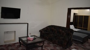 A living room with a dark patterned sofa, a glass coffee table, a flat-screen TV mounted on the wall, and a small table with a box and remote control. - Town Palace Guest House (Mekhtar)