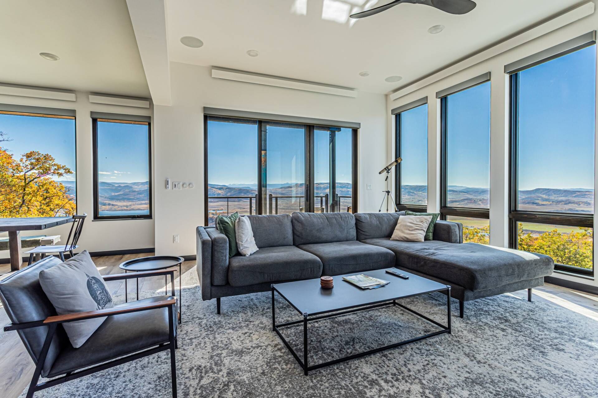 Una sala de estar moderna con un amplio sofá seccional gris, una mesa de centro y una silla. La habitación tiene ventanas grandes que ofrecen vistas a las montañas y a un árbol con hojas amarillas.