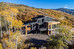 A modern multi-story house with a wooden exterior, large windows, and a balcony, surrounded by autumn foliage.