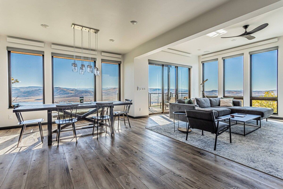 A spacious living area with a dining table and chairs, a sofa, and a large window offering a view of the mountains.