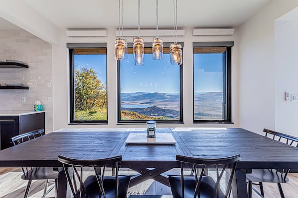 A modern dining area with a large table, chairs, and a view of the countryside through large windows.