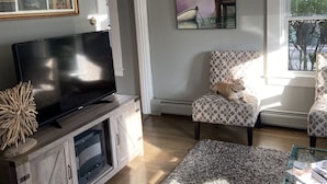 Olohuoneessa on litteänäyttöinen televisio, joka on sijoitettu puiseen kaappiin, kuviollinen nojatuoli, lasinen sohvapöytä ja seinällä oleva taulu.