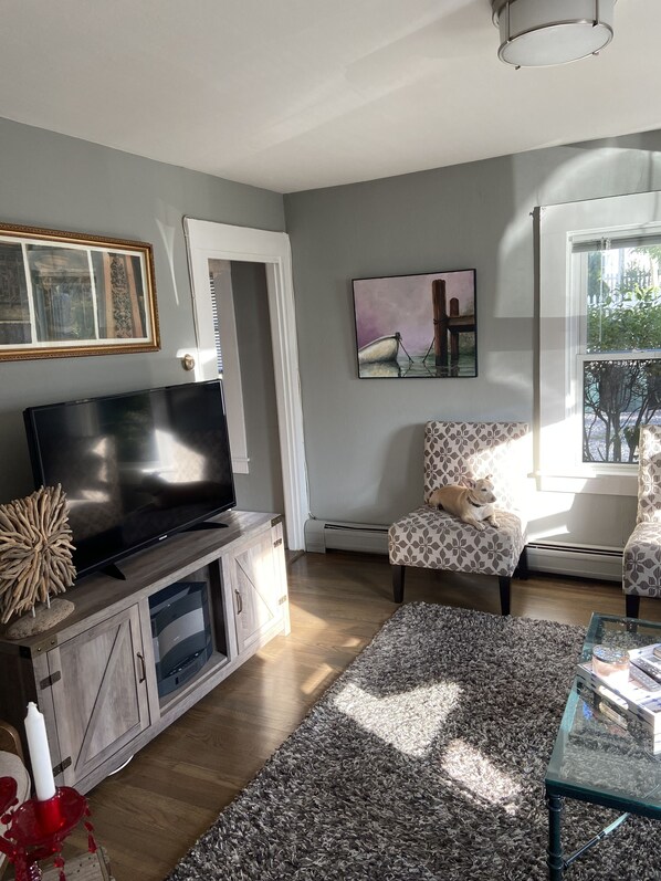A living room with a flat-screen TV on a wooden cabinet, a patterned armchair, a glass coffee table, and a painting on the wall.