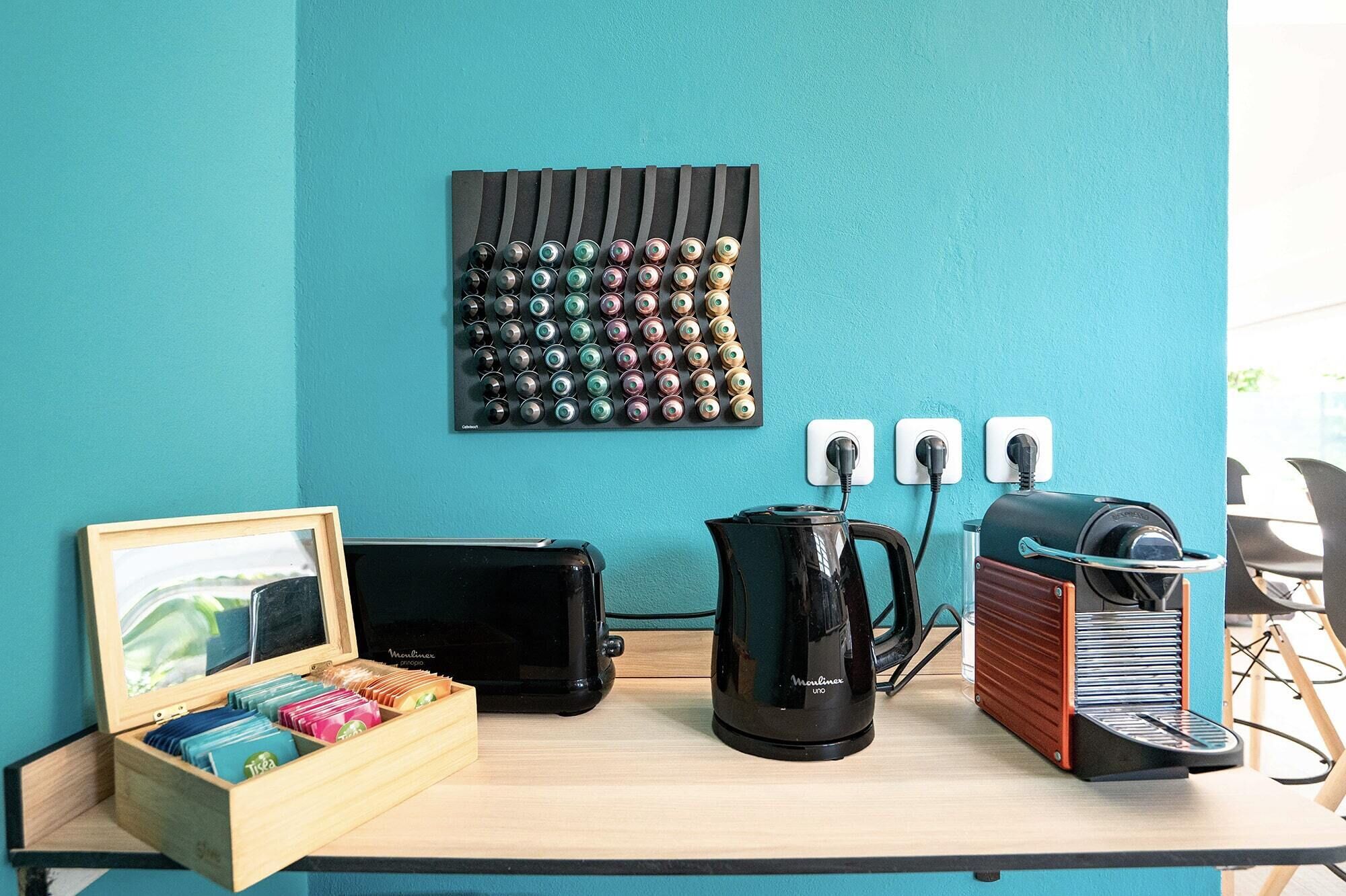 Un mur de couleur bleu sarcelle orné d’une œuvre d’art murale aux motifs noirs et bleus, une valise noire, une machine à café noire, une boîte en bois contenant des objets colorés et un appareil rouge et noir posé sur une table.