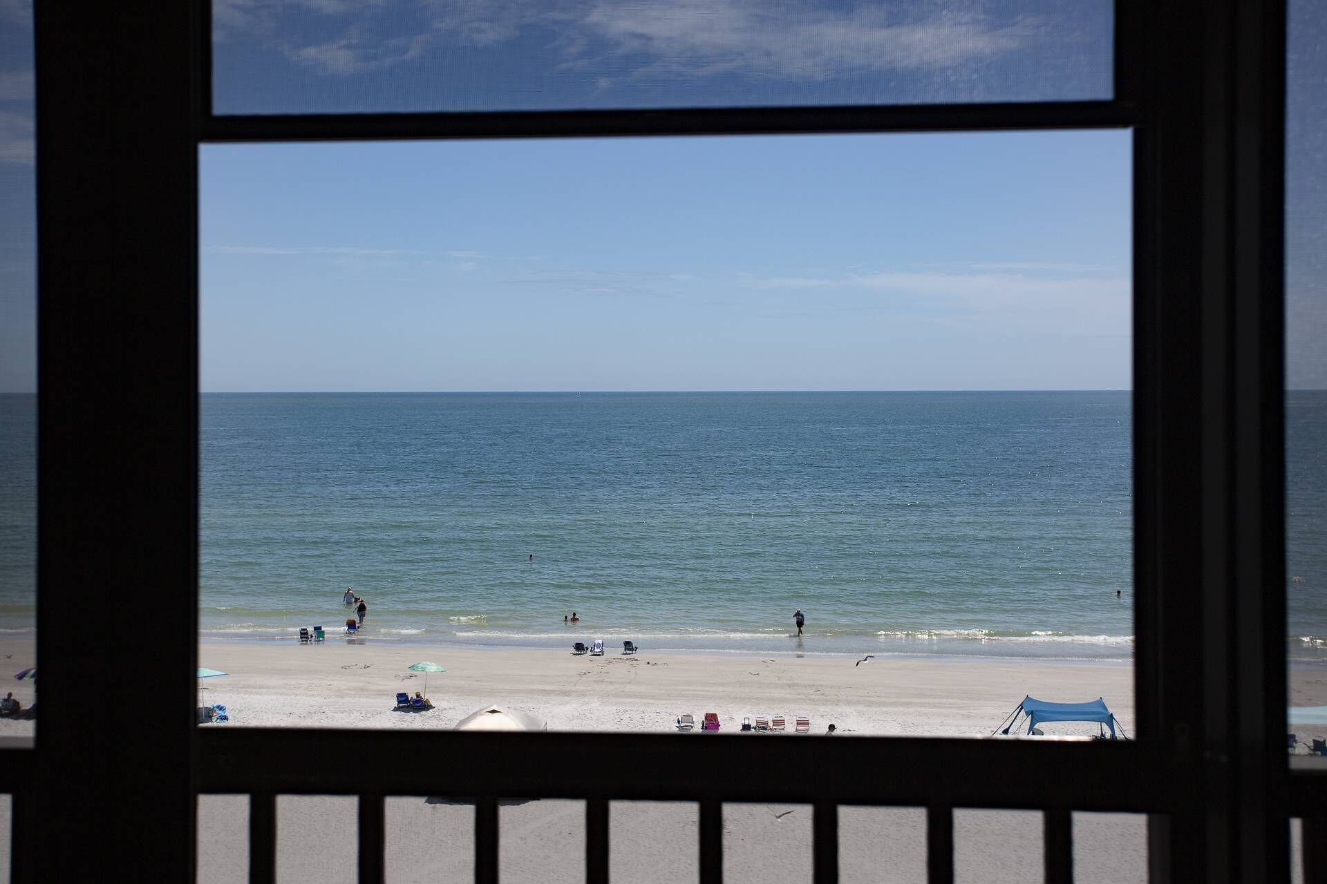 Une vue sur la plage, à travers une fenêtre ornée d’une balustrade, avec un ciel dégagé et une mer calme.