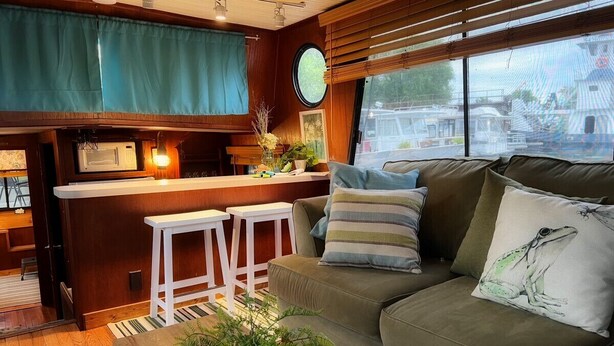 A cozy interior with a green sofa, striped pillows, a small white table, and a bar area with stools. - The "Lily Pad" Boatel Houseboat (Sabula)