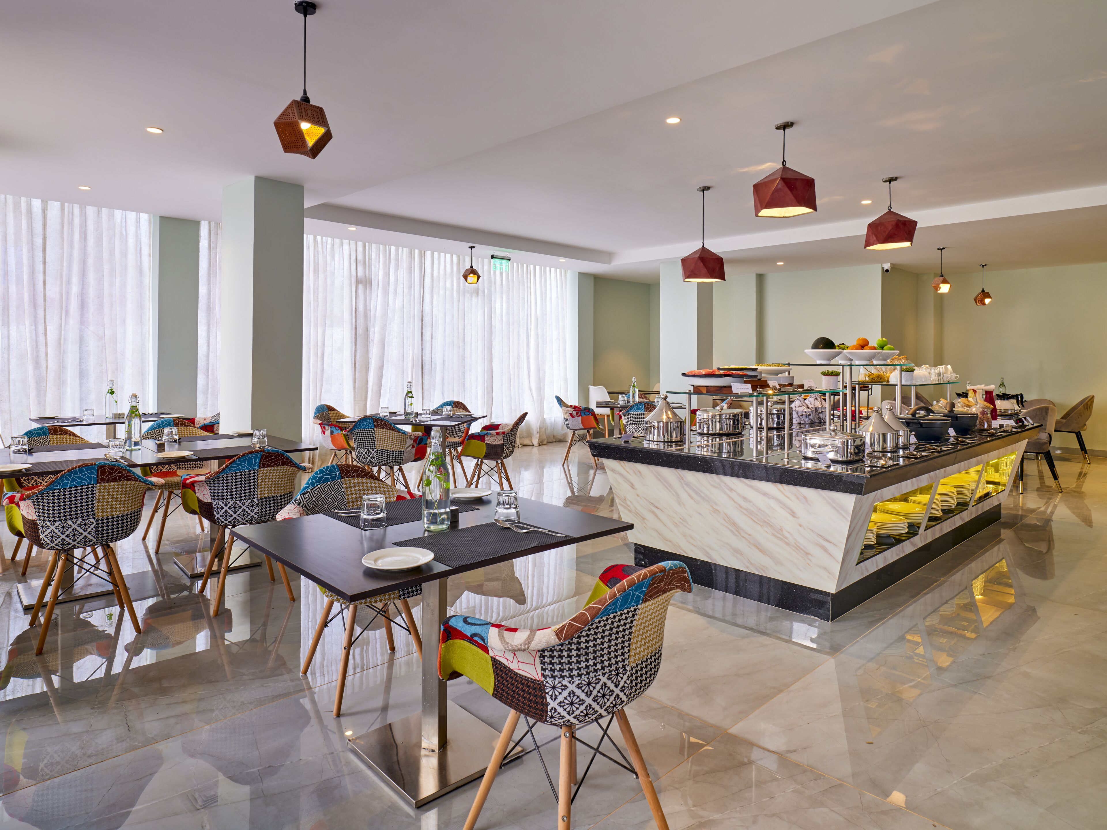 A modern hotel dining area with a central buffet, colorful chairs, and pendant lighting.