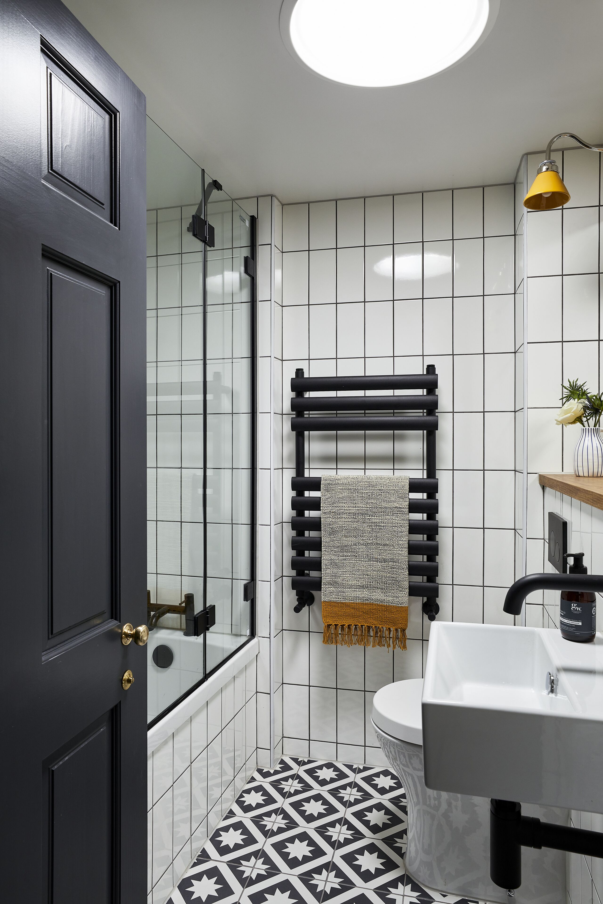 A bathroom with a black door, white tiled walls, a black and white patterned floor, a white sink, and a yellow light fixture.
