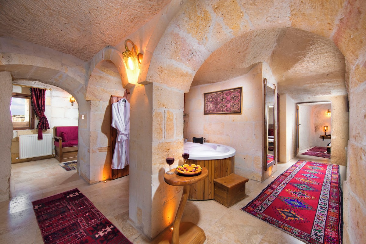 A stone-walled bathroom with a bathtub, a wooden bench, and a fruit bowl with a glass of wine.