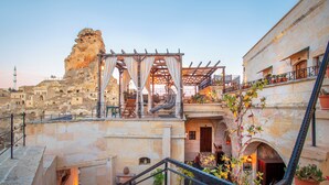 Une cour dotée d’un escalier en pierre, d’une pergola en bois et offrant une vue sur un bâtiment historique ainsi que sur une formation rocheuse.