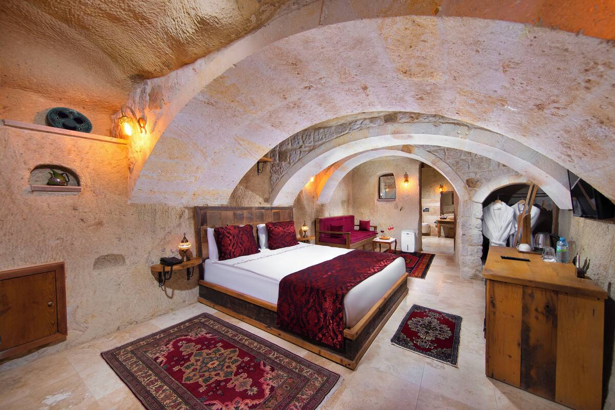 A bedroom with a stone ceiling, a bed with red bedding, a wooden dresser, and a small table with a lamp.