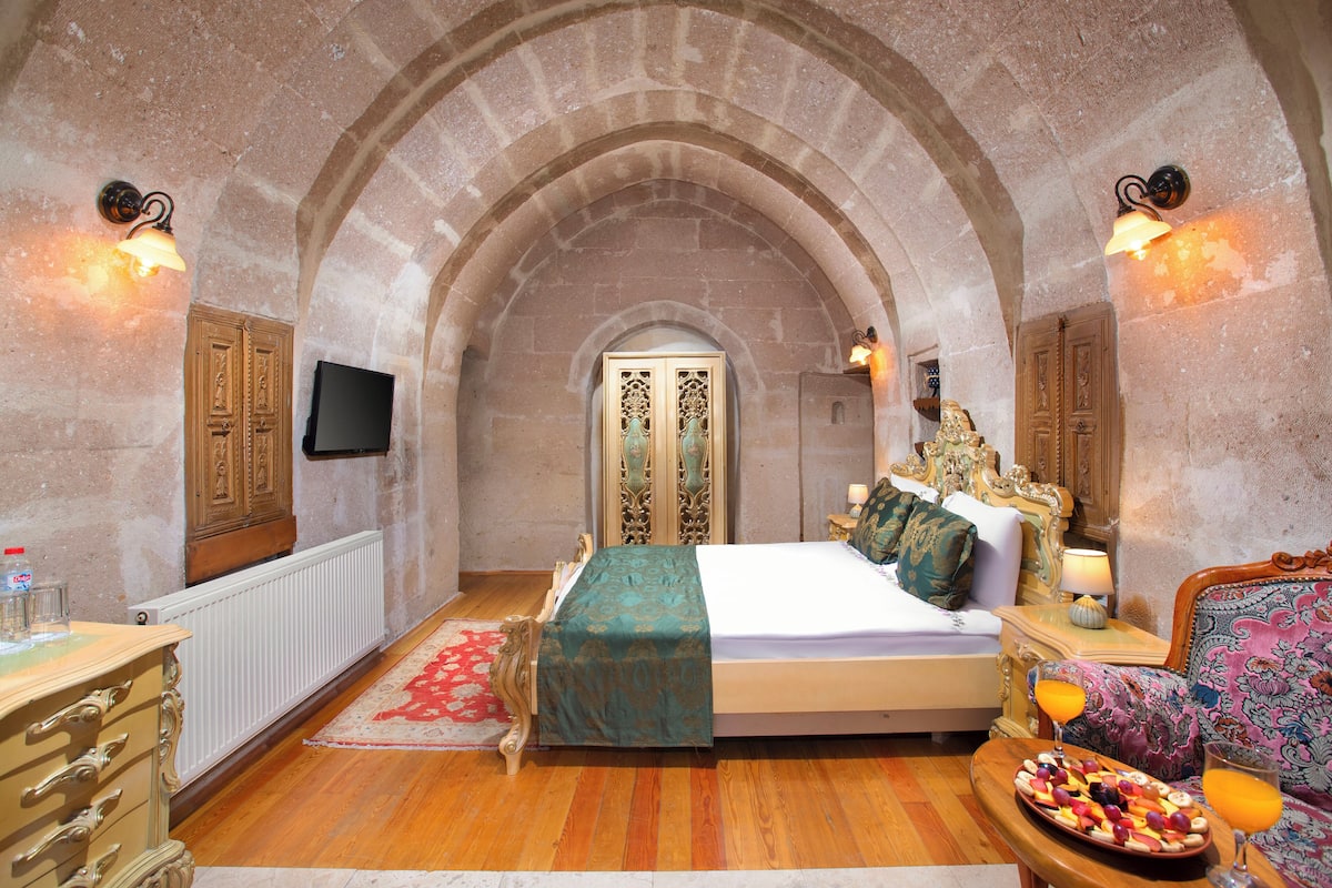 A bedroom with an arched ceiling, a bed with a green bedspread, a wooden dresser, a nightstand with a lamp, and a fruit platter on the side table.