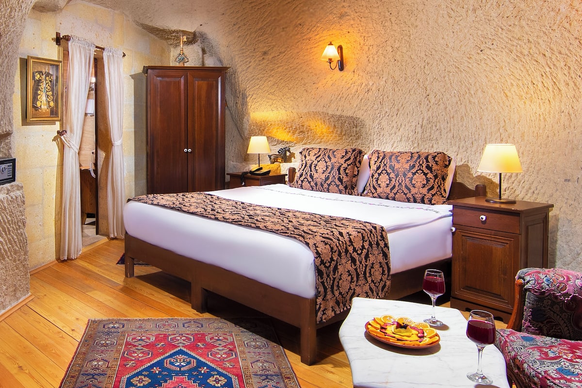 A bedroom with a large bed, wooden wardrobe, bedside lamps, a dining table with a fruit platter and wine glasses, and a patterned rug.