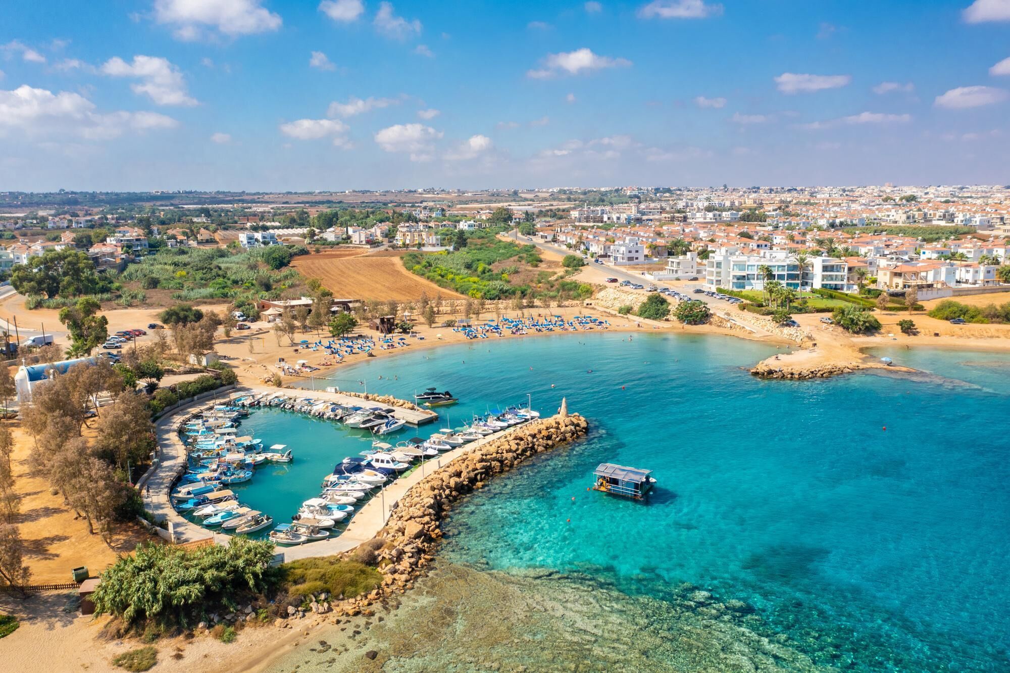 Marina costeira com barcos atracados, uma praia arenosa com guarda-sóis e área residencial ao fundo.