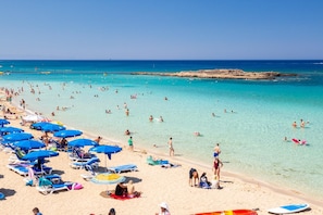 Une plage bondée où de nombreuses personnes se baignent et prennent des bains de soleil. Des parasols et des chaises longues sont disposés le long du rivage.