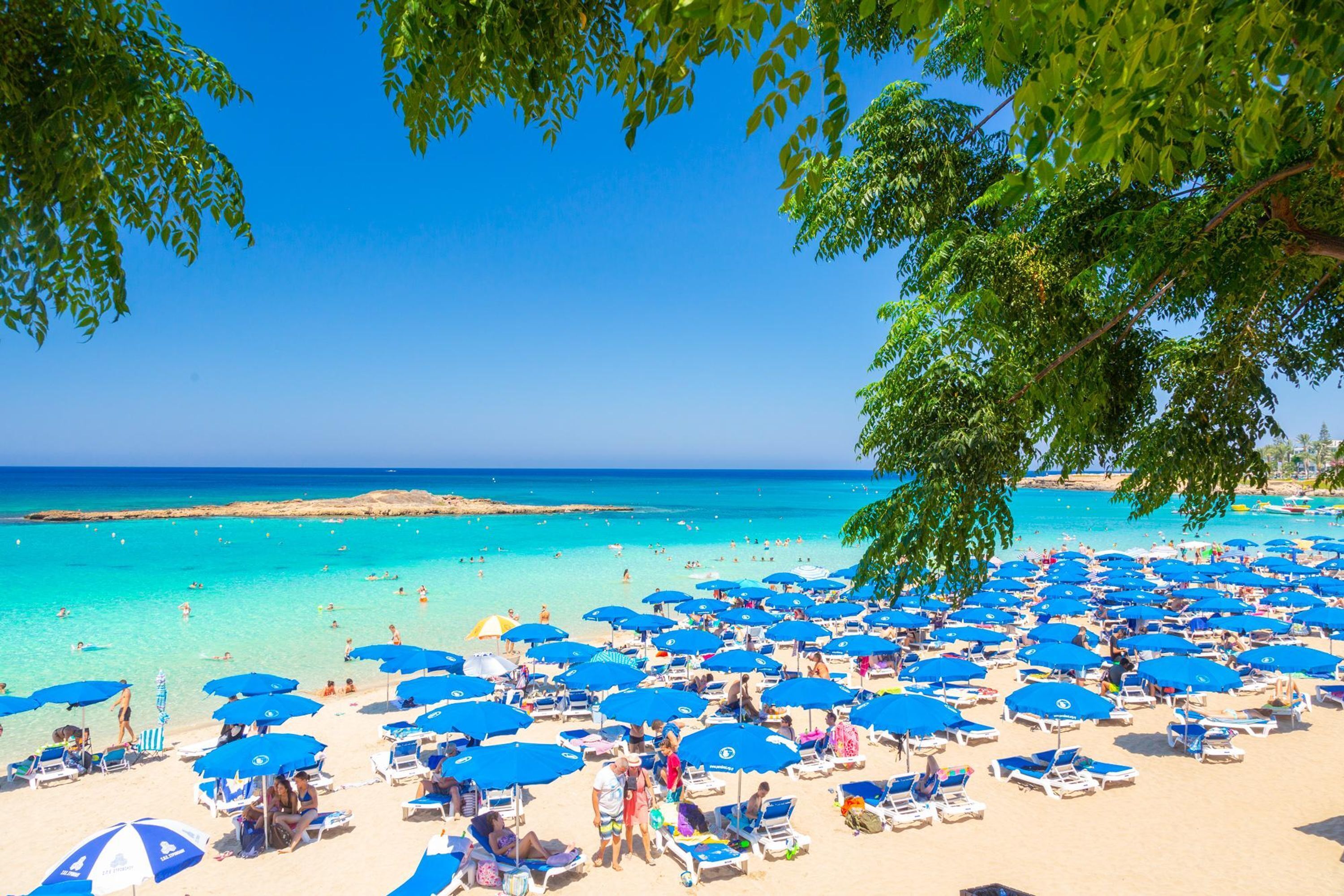 A beach with numerous blue umbrellas and sun loungers, clear turquoise water, and a clear sky.