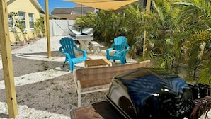A patio area with outdoor furniture, including two blue Adirondack chairs and a wicker table, under a large shade sail. - Book & Stay Today + King Bed + Beach Chairs (Indian Rocks Beach)