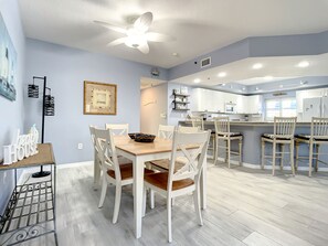 Una cocina moderna con armarios blancos, una isla central y una zona de comedor con sillas blancas y una mesa de madera.
