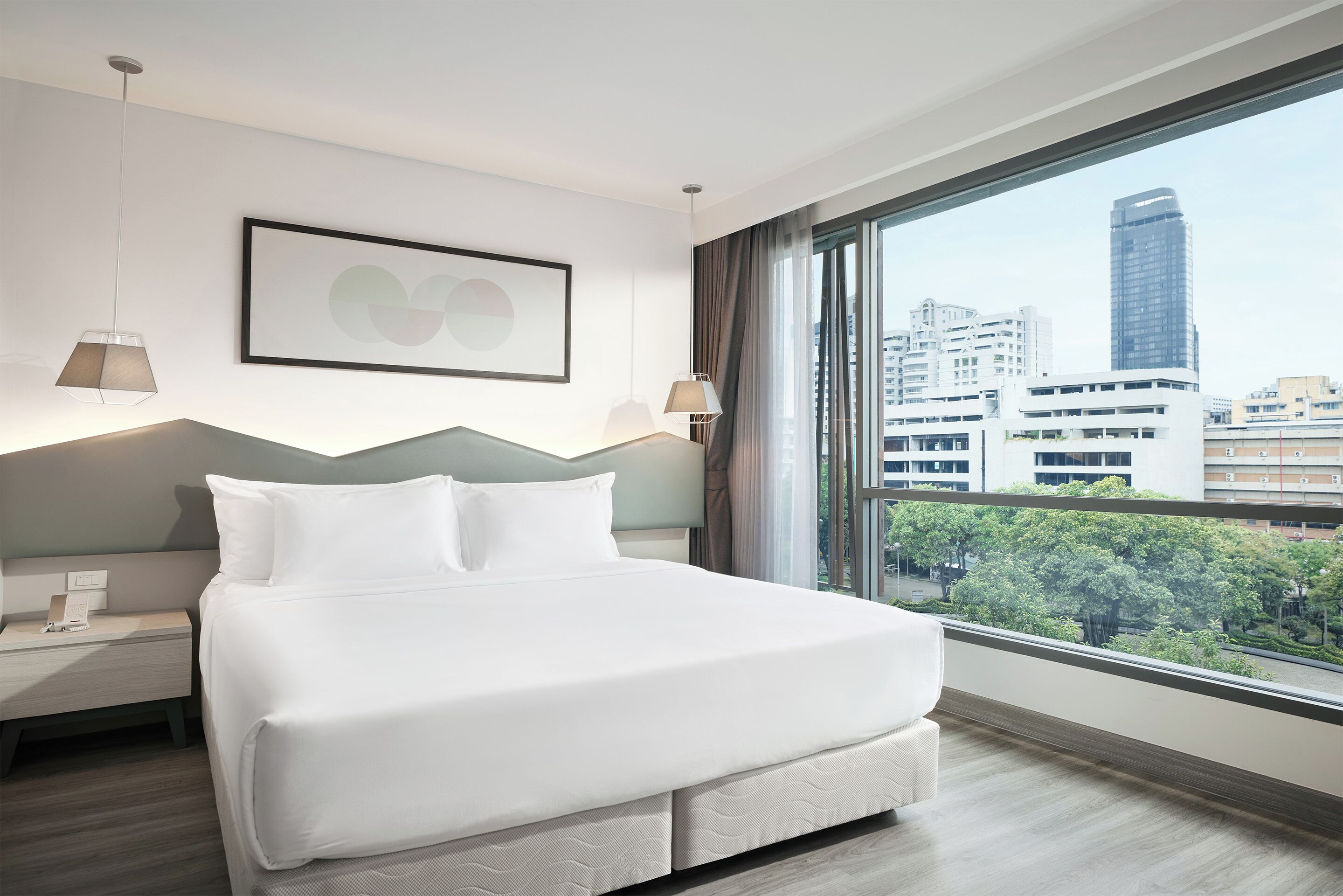 A modern hotel room with a large bed, two bedside tables, a framed artwork, and a city view through large windows.