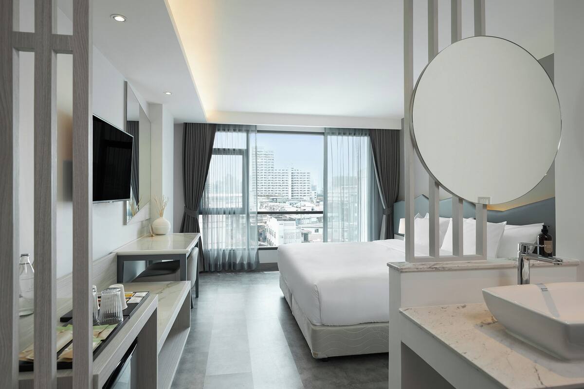 Una habitación de hotel moderna con una cama grande, un espejo redondo, un tocador con lavamanos y un escritorio con televisor de pantalla plana.