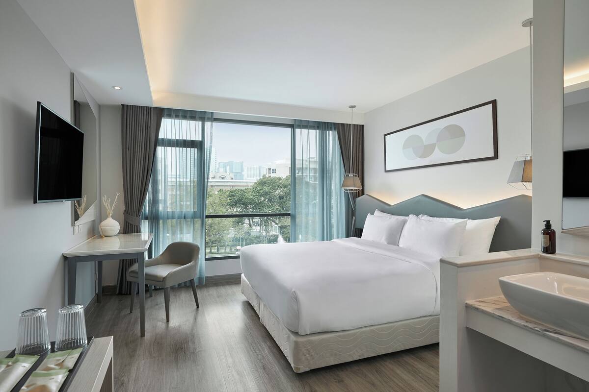 Habitación de hotel moderna con una cama grande, un escritorio, una silla, un televisor, un baño con lavamanos y vistas a la ciudad.
