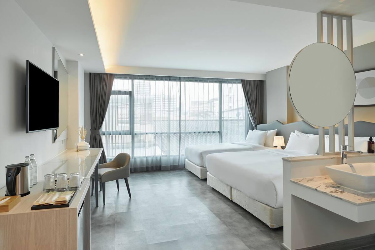 Una habitación de hotel moderna con una cama grande, un escritorio con televisor de pantalla plana, un baño con espejo amplio y lavabo, y vistas de la ciudad a través de ventanas grandes.